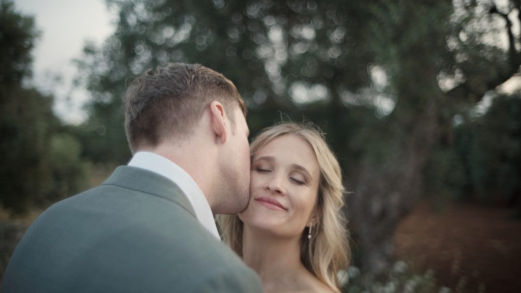 the groom kisses the bride who smiles with her eyes closed among the olive trees at sunset