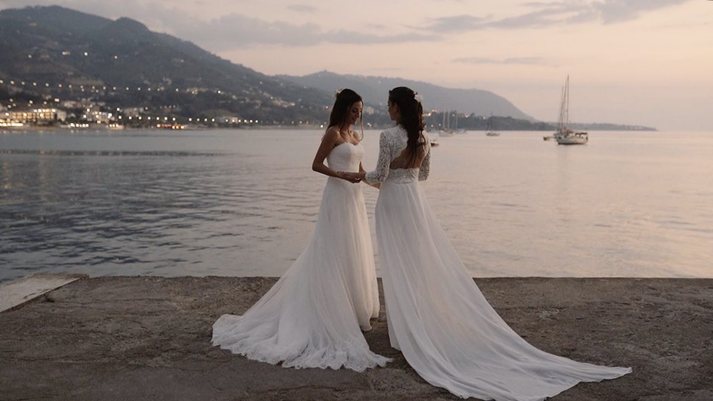 two brides in white dresses look at each other holding hands on the port of Cefalù in Sicily at sunset