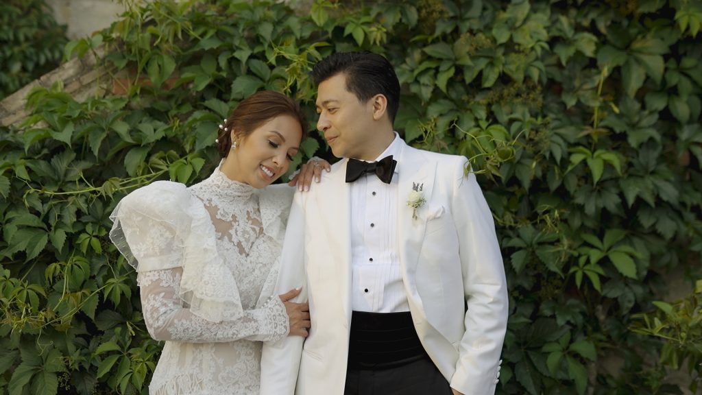 bride leaning on the groom's shoulder smiles. They are in front of a green wall of leaves