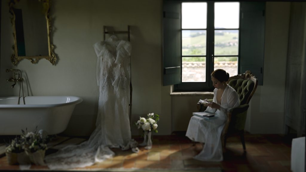 the bride goes over her vows  before her destination wedding sitting on an armchair in front of a window with her wedding dress and bouquet next to her
